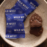 Two Wild West Huckleberry & Cashew Bear Claws nut cluster wrappers next to a chocolate bear paw treat, arranged on a speckled beige ceramic plate.